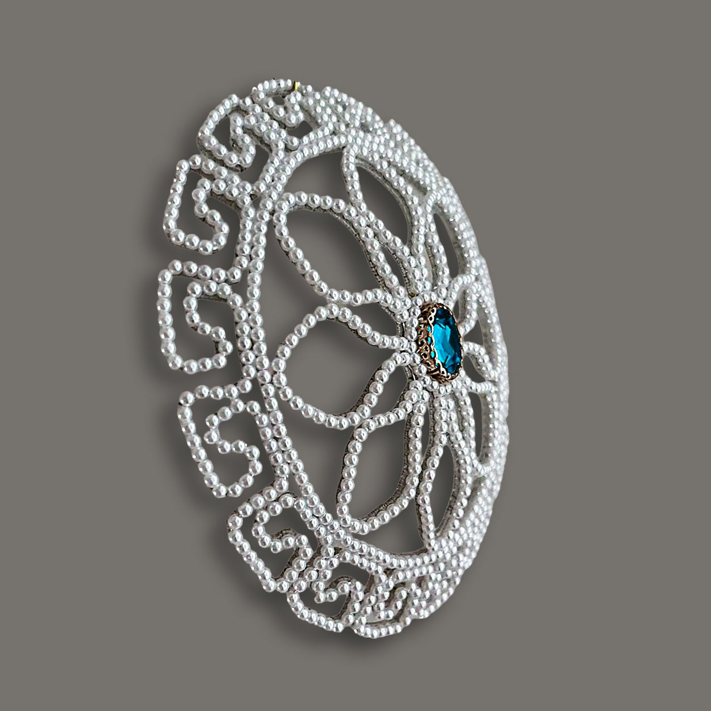 Angled view of a domed pearl bridal bun cover featuring a unique geometric Greek key outer border, floral inner beadwork, and a blue sapphire crystal center on a grey background.