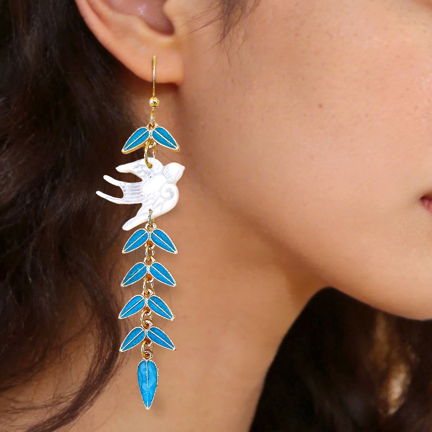 Detailed close up of a delicate white shell swallow and blue leafy vine earring worn on the earlobe