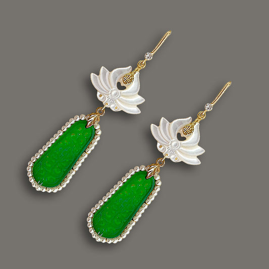Angled view of elegant carved white shell lotus earrings with vivid green stone drops and pearl borders on dark background