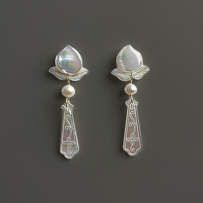 A pair of elegant bridal stud earrings featuring hand-carved white mother of pearl flowers and freshwater pearl drops against a dark taupe background.