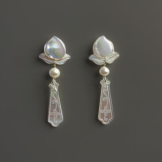 A pair of elegant bridal stud earrings featuring hand-carved white mother of pearl flowers and freshwater pearl drops against a dark taupe background.